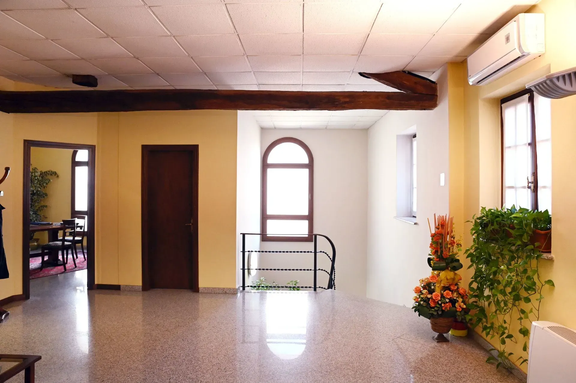 Stanza interna con pareti e pavimenti chiari, dotata di porta, finestra ad arco e piante sospese.