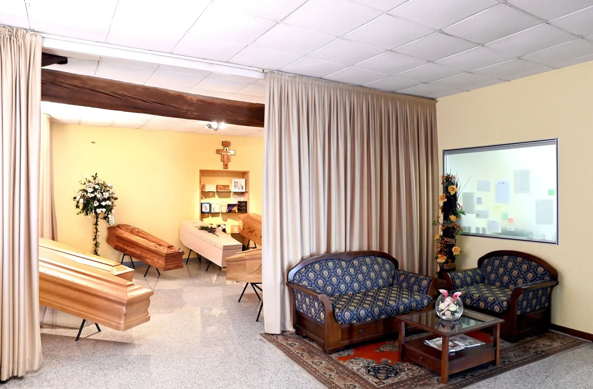 Interno di un'agenzia di pompe funebri con bare, area salotto e una croce su una parete.