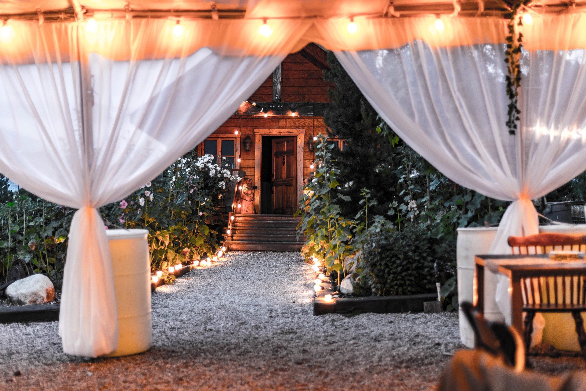 A tent with white curtains and lights is sitting in the dirt in front of a house.