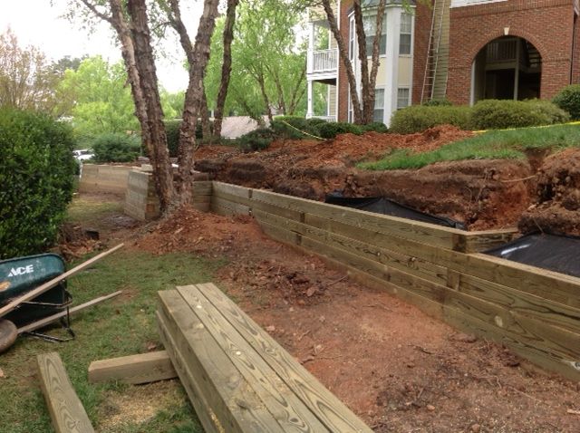 Wooden retaining wall under construction next to a building and greenery.