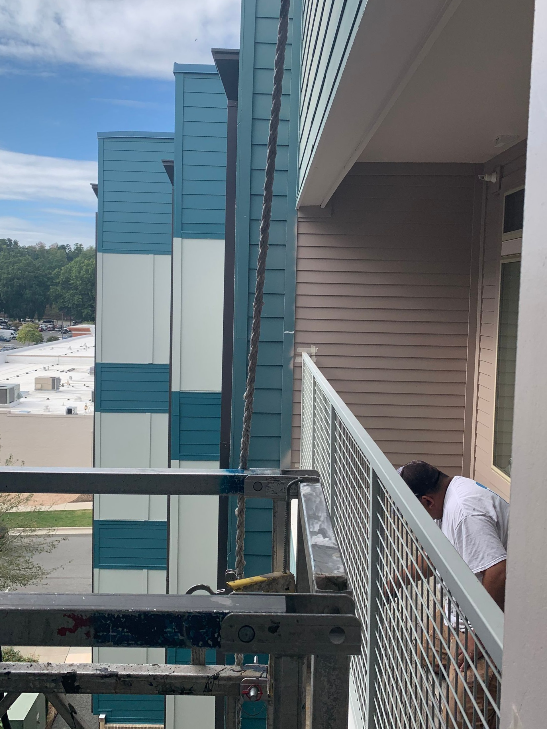 Building exterior with blue, white, and teal panels. A person stands on a balcony. Scaffolding is below.