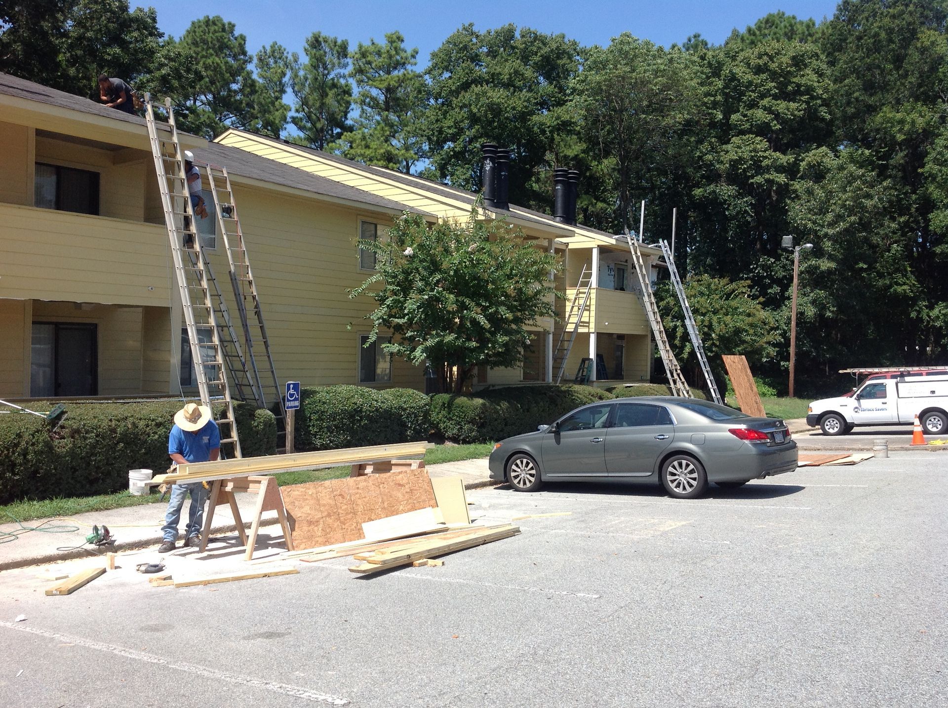 Workers on ladders repairing a light yellow apartment building roof. Gray car and white truck parked nearby. Sunny day.