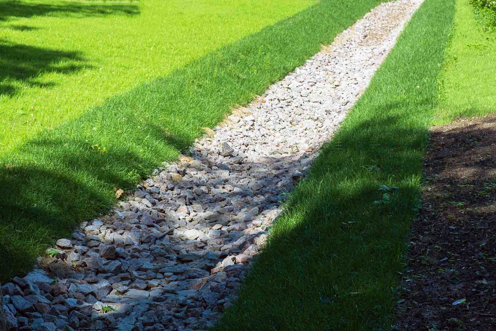 Gravel-filled drainage ditch running through green lawn, bordered by grass.