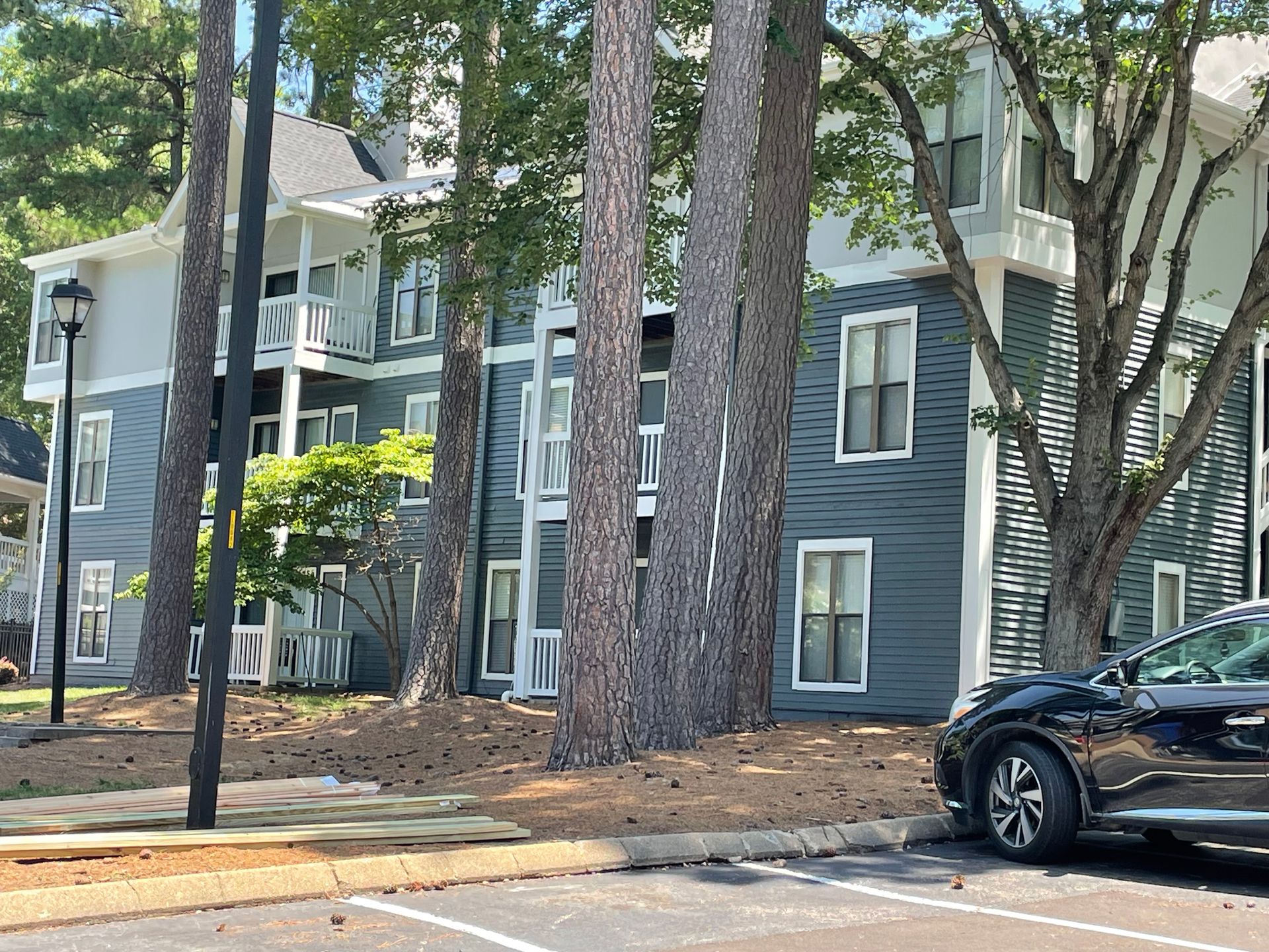 Apartment building with blue siding and white trim, surrounded by trees. A car is parked nearby.