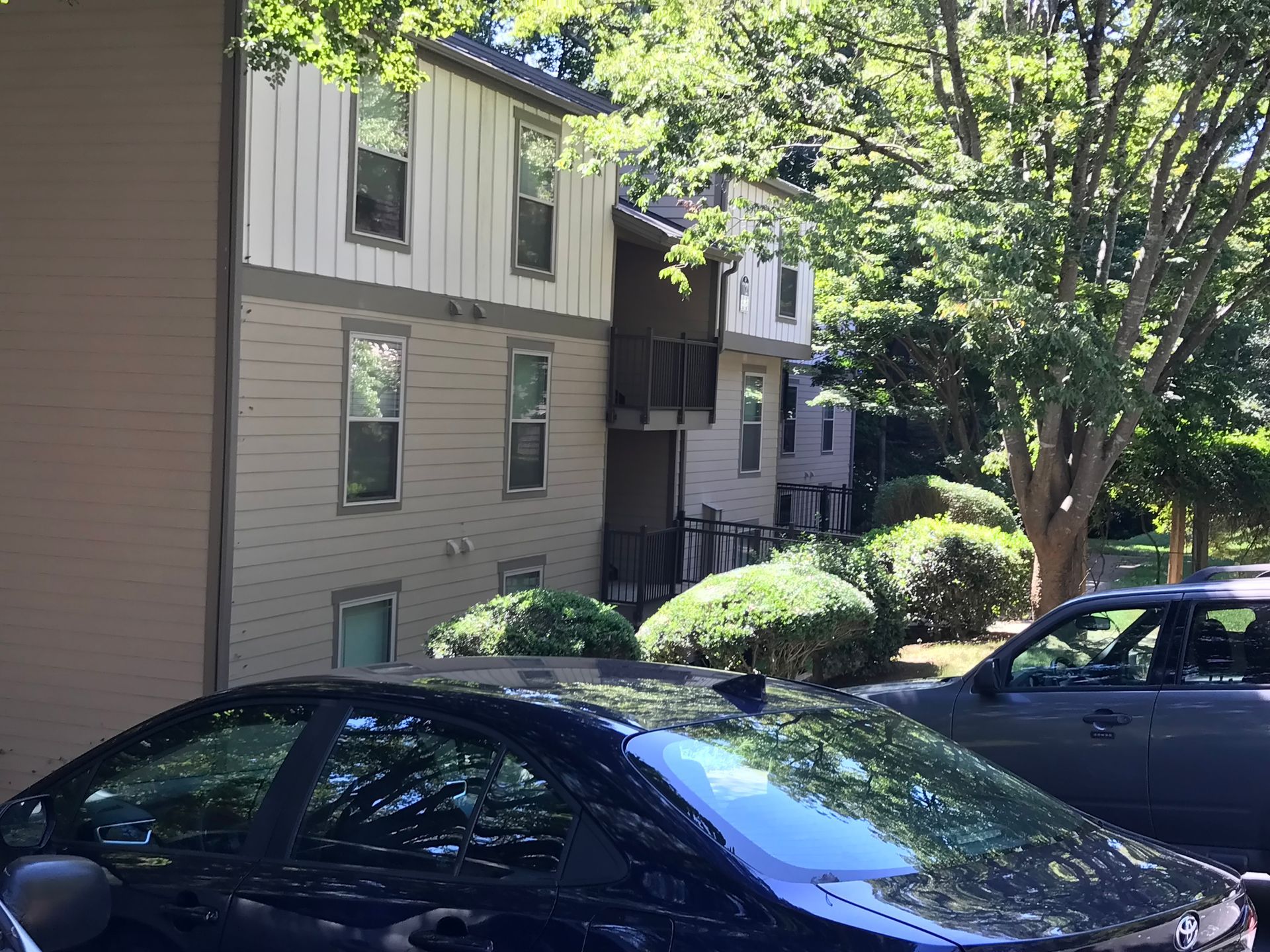 Apartment building exterior with cars parked in front, trees, and blue sky.