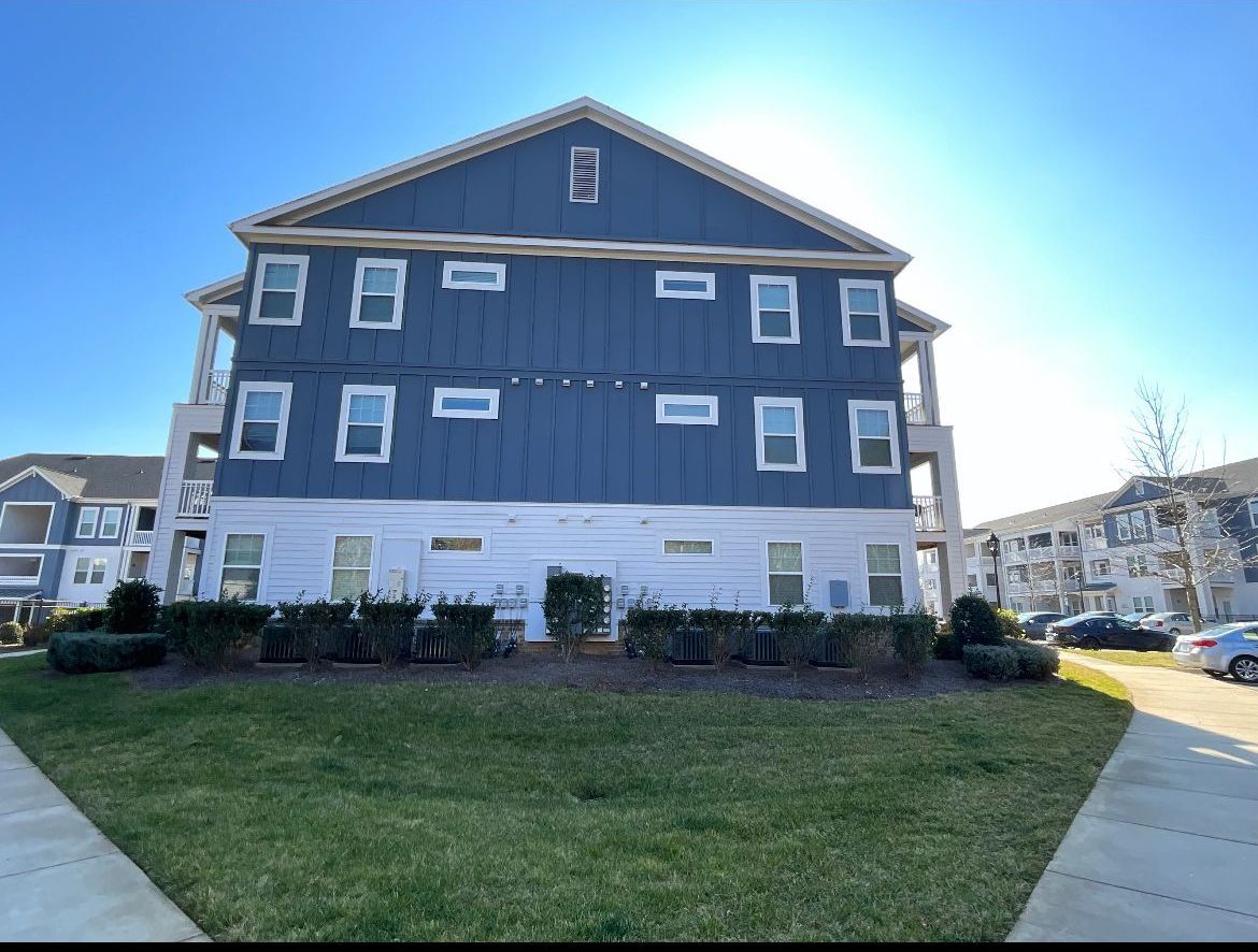 Blue and white apartment building with multiple windows, on a sunny day. Green grass and sidewalk in the foreground.