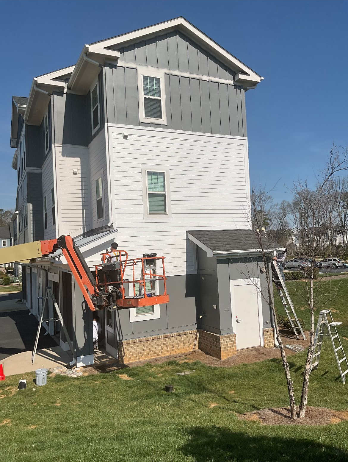 Construction on multi-story building. A lift and ladders are used for siding installation; some walls are painted gray.