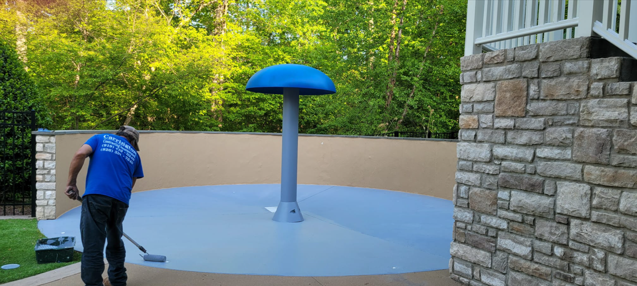 A person painting a blue circle around a mushroom-shaped water feature in an outdoor area.