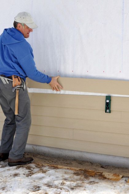 A person installing beige siding on a building with white wrap; setting is outdoors, likely winter.