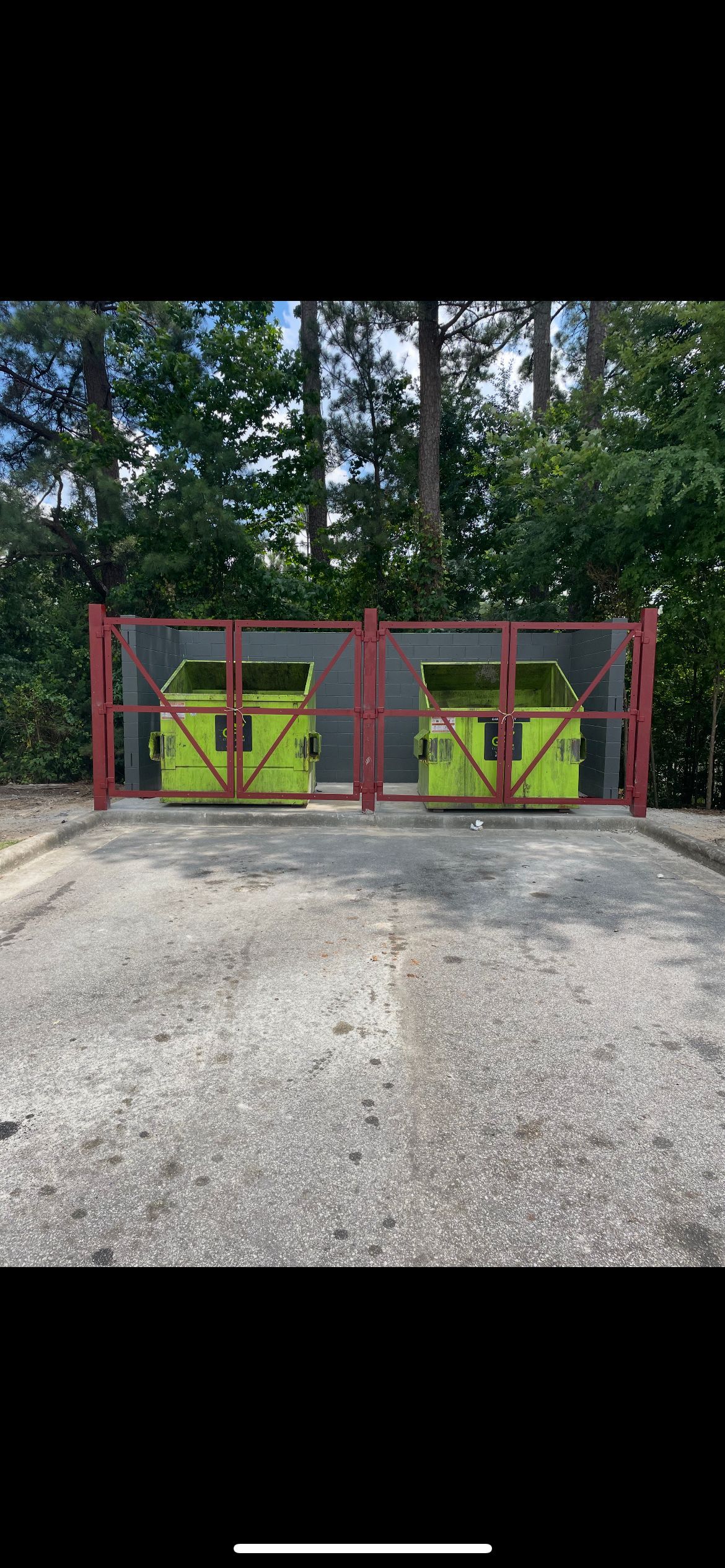Two yellow dumpsters behind a metal gate on a gravel surface, surrounded by green trees.