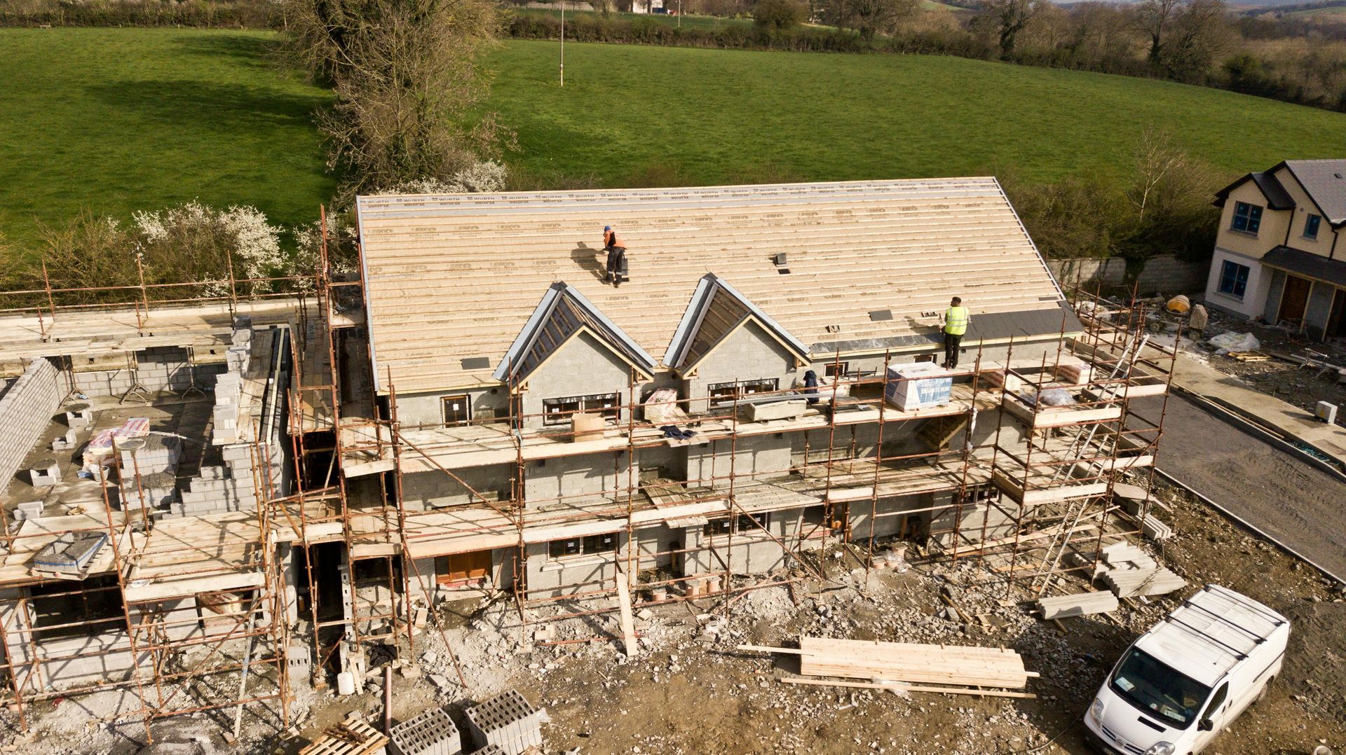 Construction site, partially built house with workers on the roof. Gray stone, green grass background, white van.