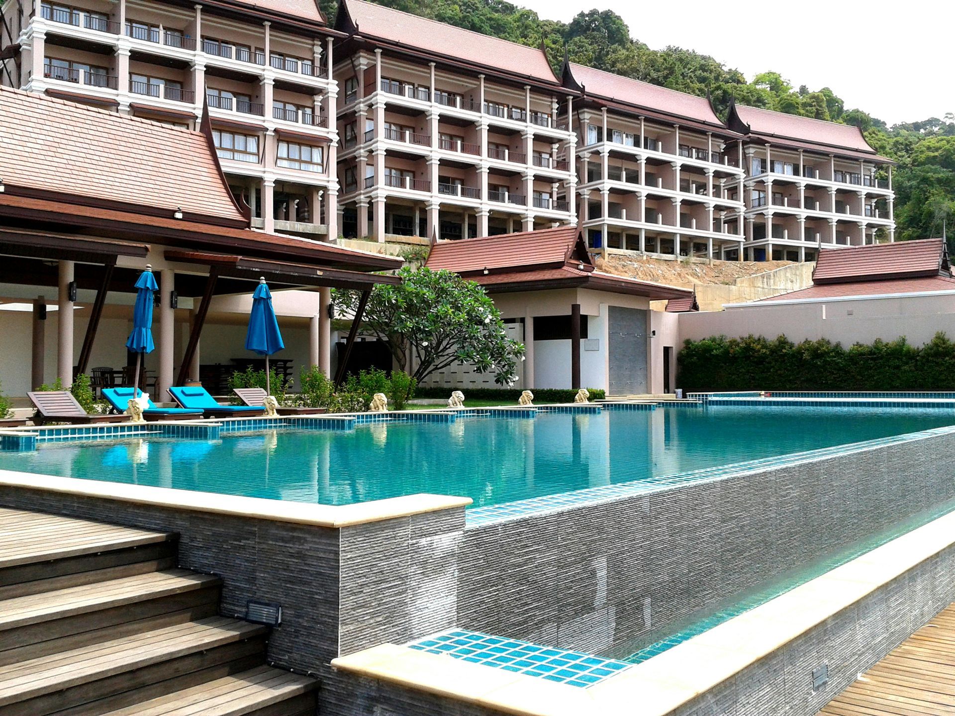 Swimming pool in front of a multi-story building. Blue umbrellas and lounge chairs near pool.