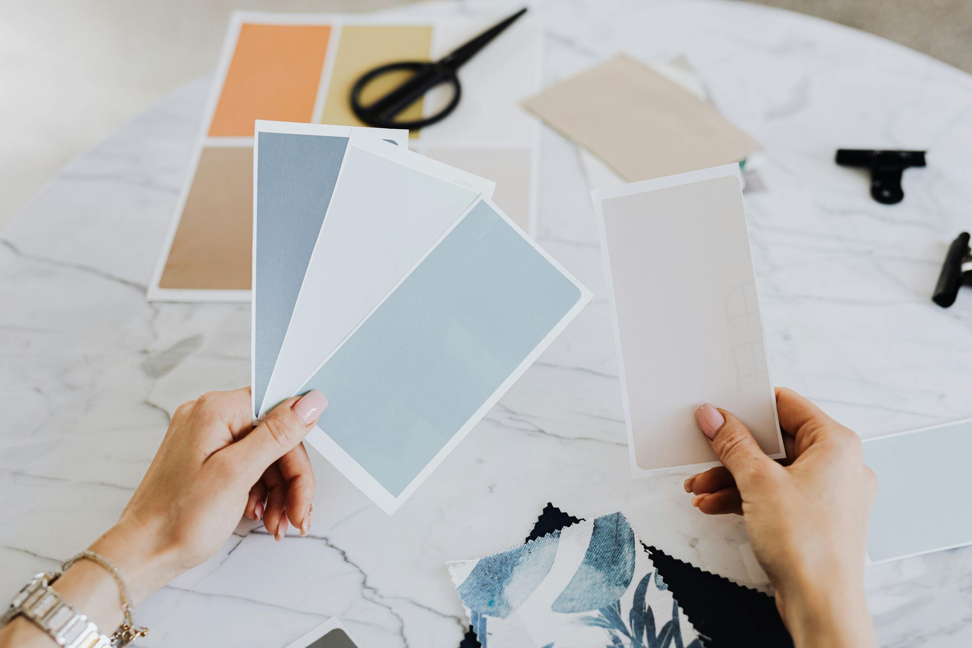 Hands holding paint swatches on a marble table with scissors and other swatches.