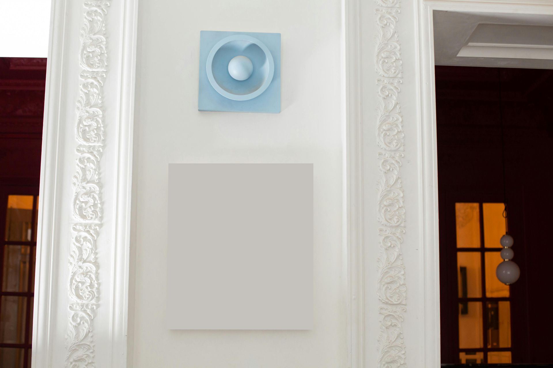 White wall with square painting below a light blue abstract sculpture, framed by ornate white molding.