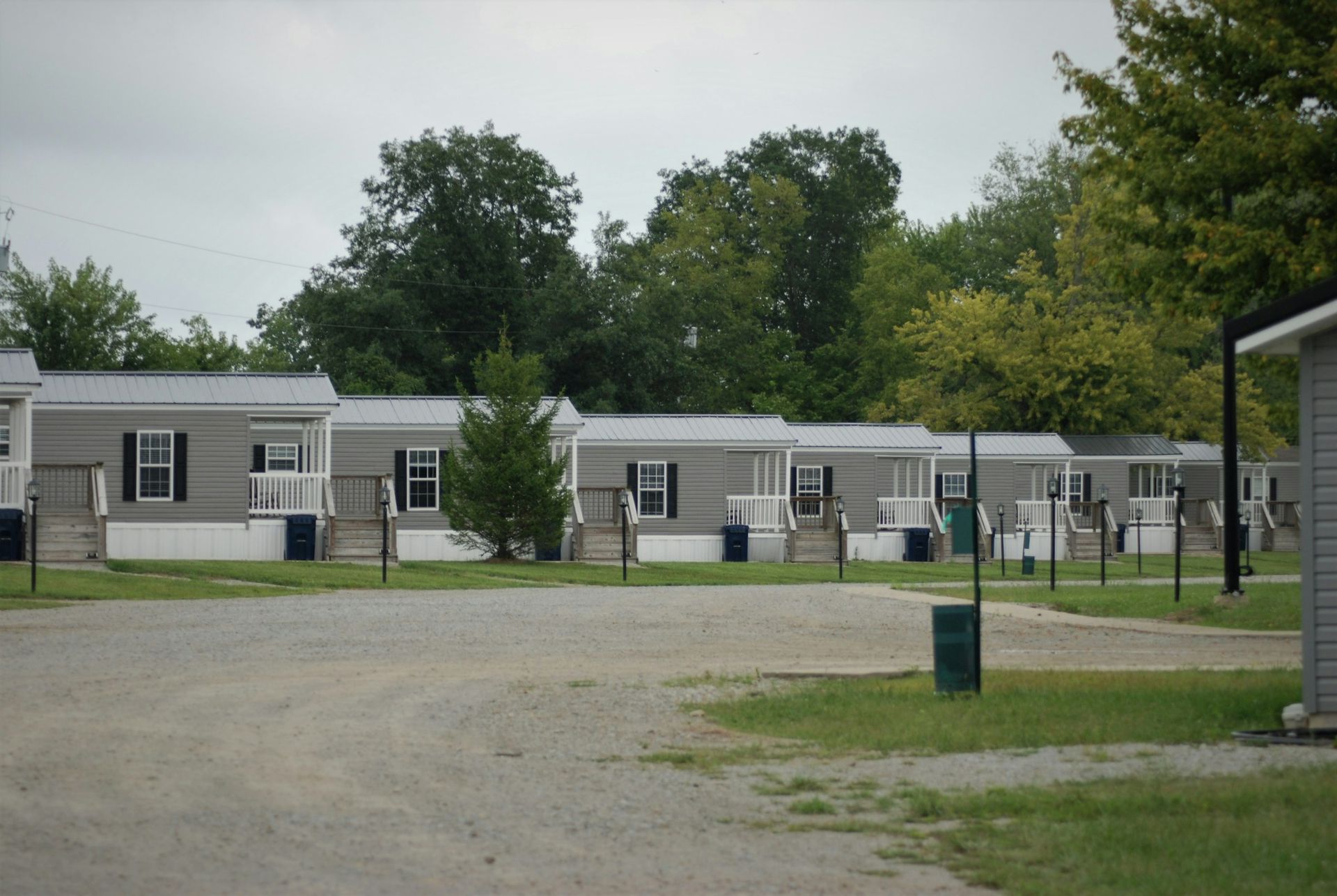 Gray mobile homes in a row, gravel parking area, green grass and trees. Cloudy sky.