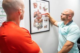 A person points to an anatomical digestive system chart on a wall while consulting with a person in an orange shirt.