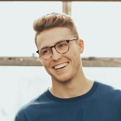 Een jonge man met een bril en een blauw shirt lacht.