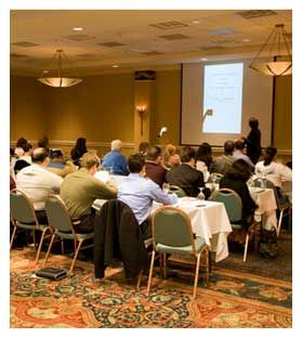 A group of people are sitting at tables in a conference room watching a presentation.