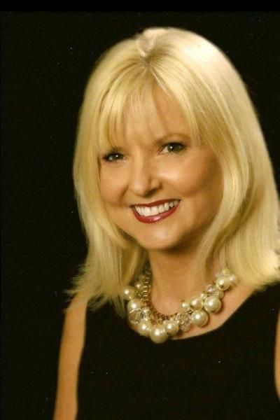 A woman with blonde hair and a pearl necklace smiles for the camera