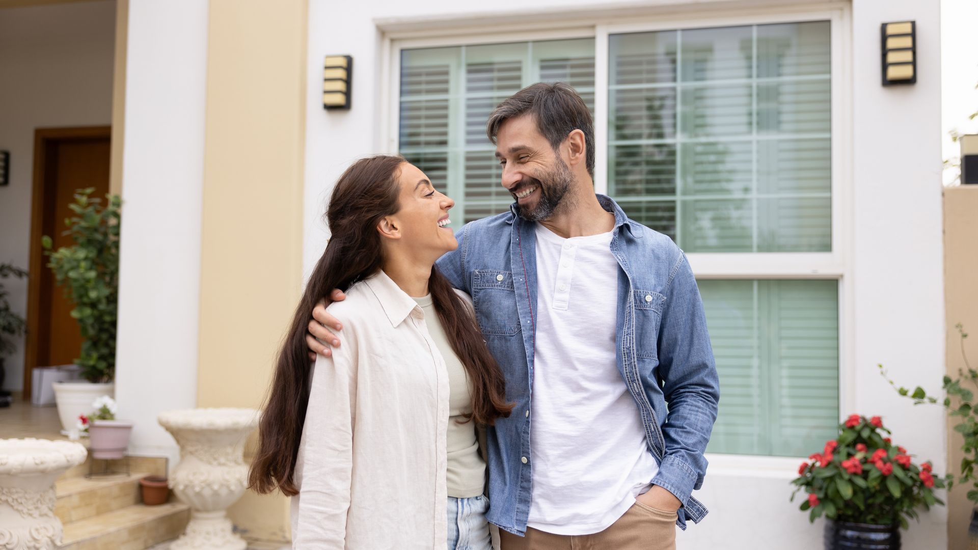 A couple standing together in front of their home, smiling as they look at each other.