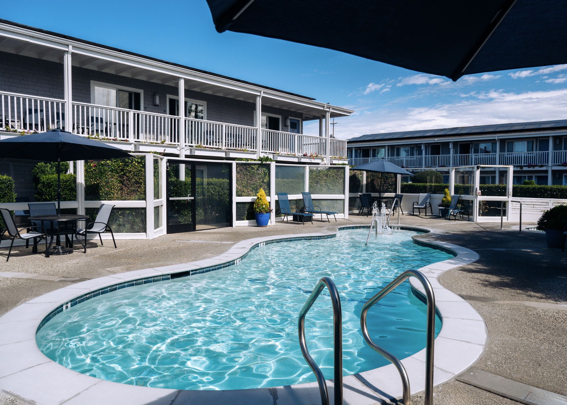 A sunlit outdoor swimming pool with a central fountain, surrounded by lounge chairs and a multi-story building.
