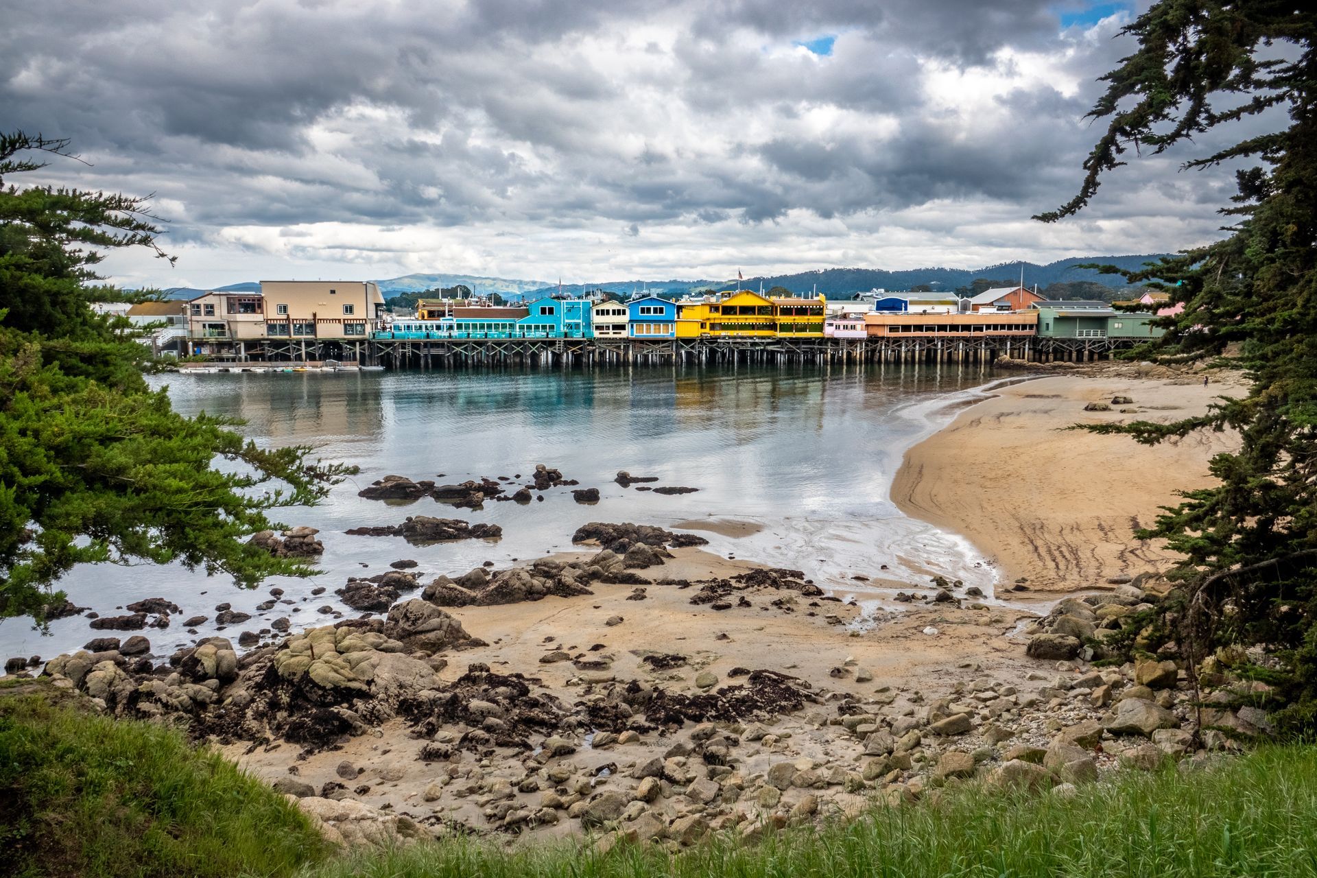 Cloudy harbor with colorful waterfront buildings, rocky shore, and a sandy beach beside calm water.