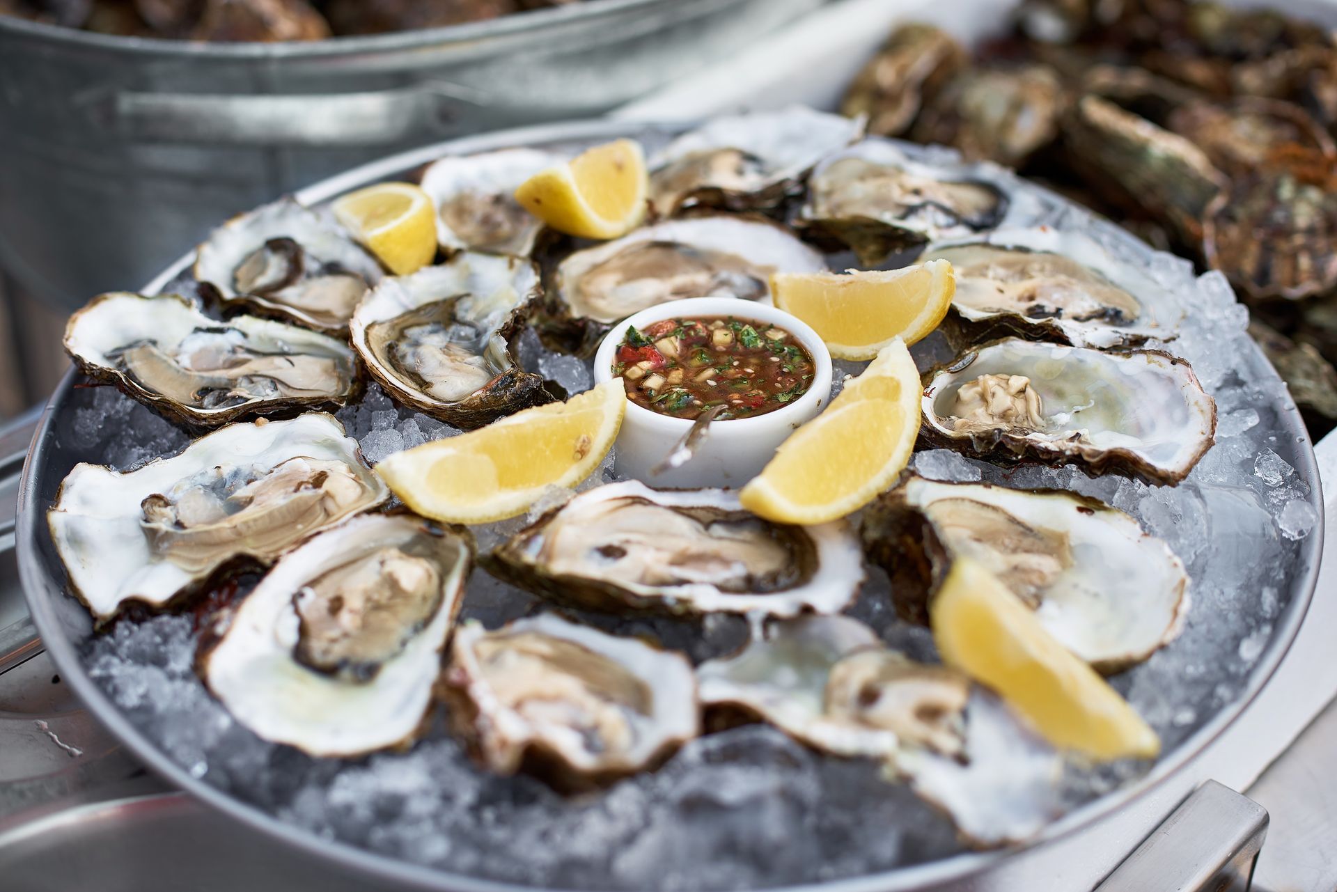 A platter of raw oysters on crushed ice with lemon wedges and a small bowl of dipping sauce.