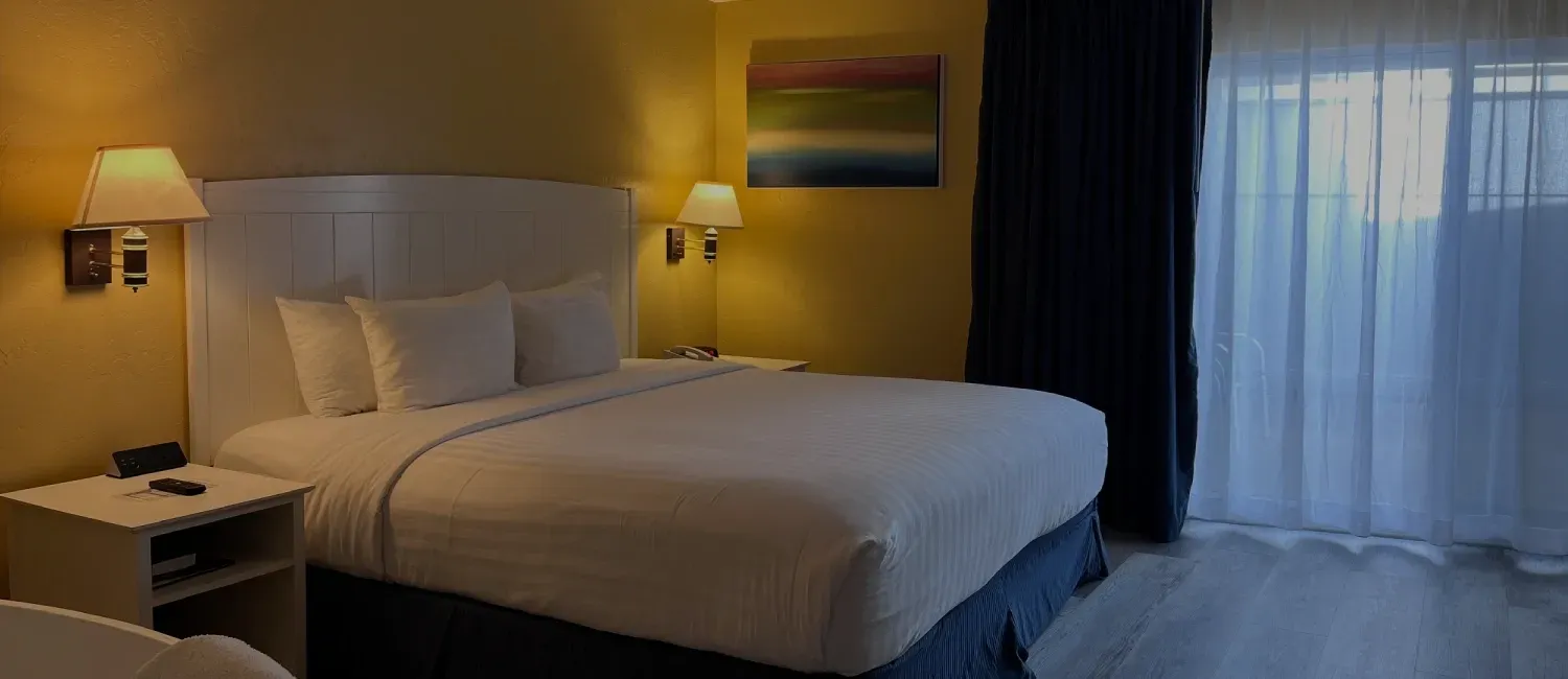 A hotel room with a neatly made bed, white linens, yellow walls, bedside lamps, and floor-to-ceiling curtains.