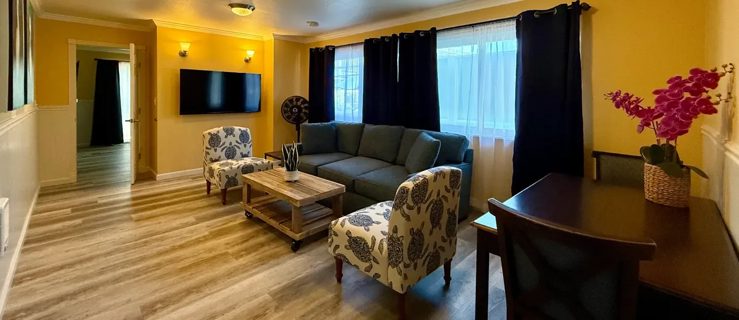 A living room with yellow walls, a grey sectional, patterned armchairs, a television, and a dining table with flowers.
