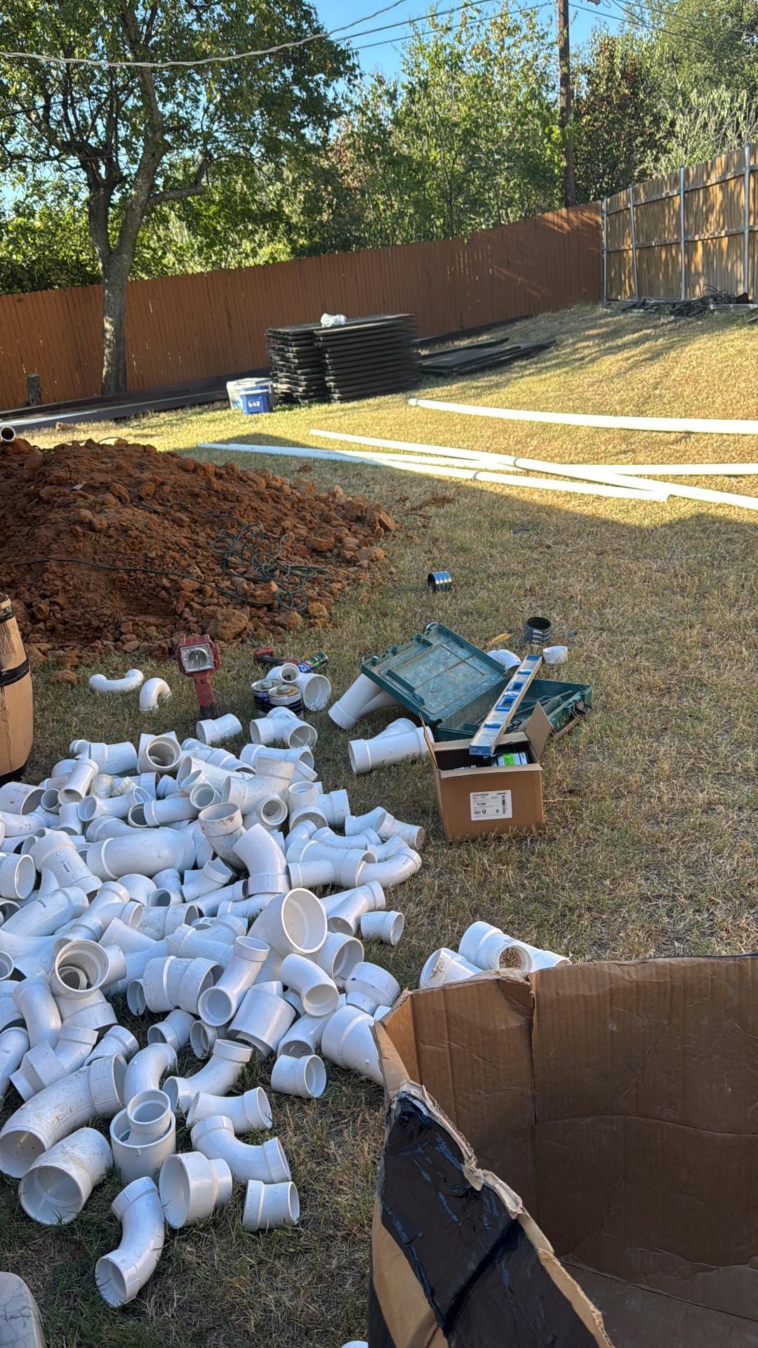 Construction site with scattered white PVC pipes, a pile of dirt, and a cardboard box on grass.