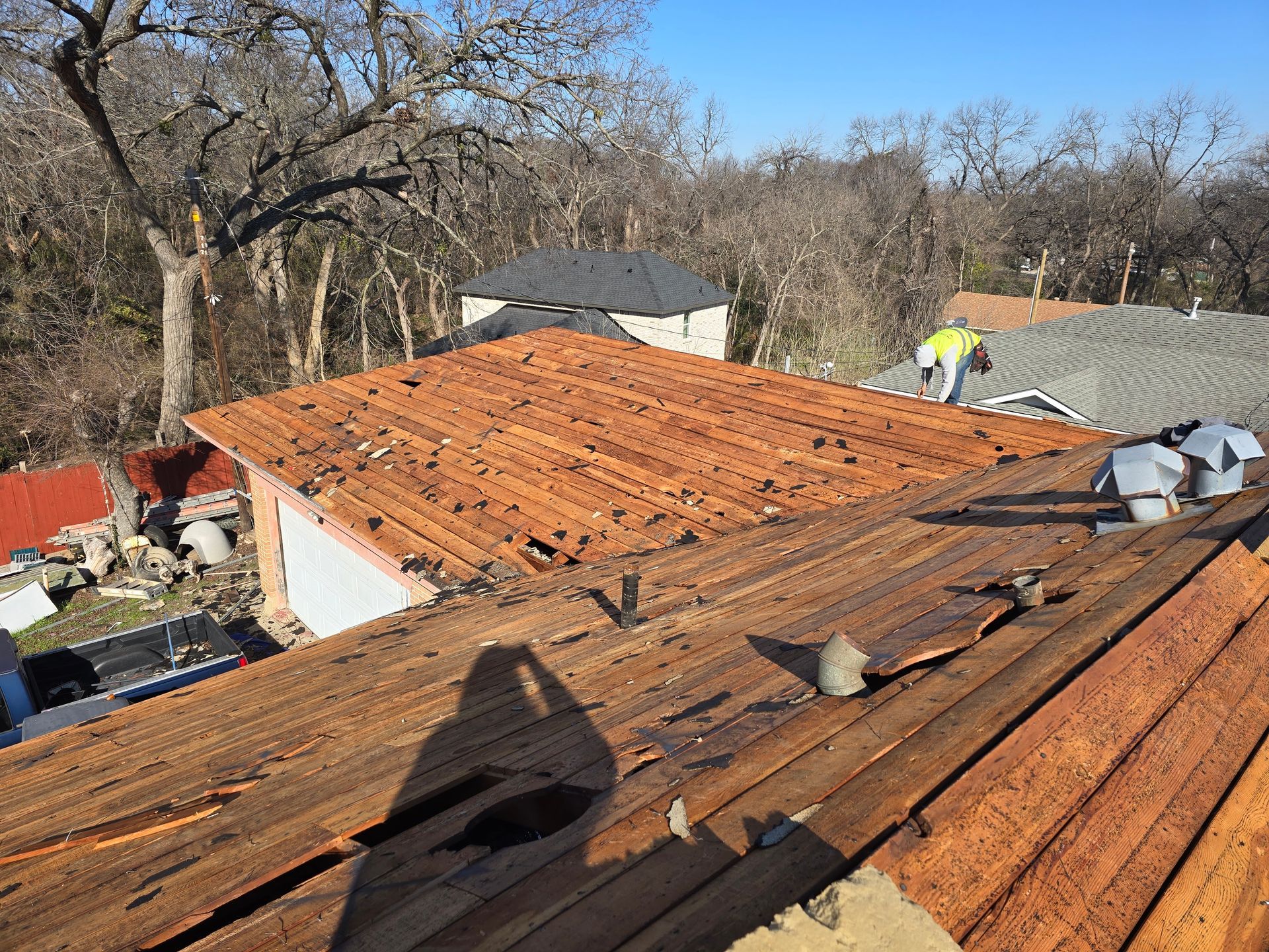 Damaged wooden roof with missing shingles; person working on the roof. Sunny day.
