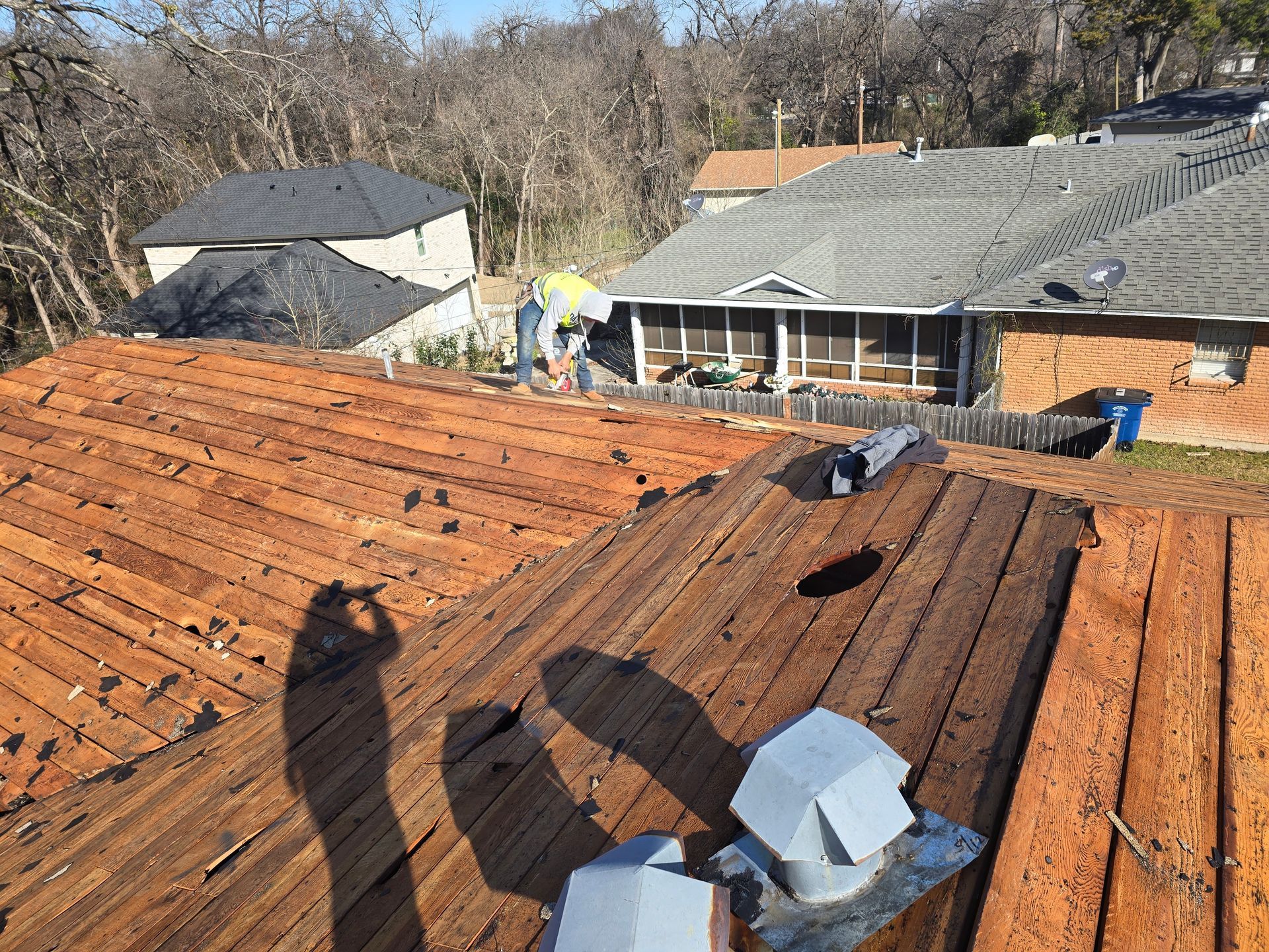 Roofer repairs an old, weathered roof. Other houses and trees are in the background. Sunny day.