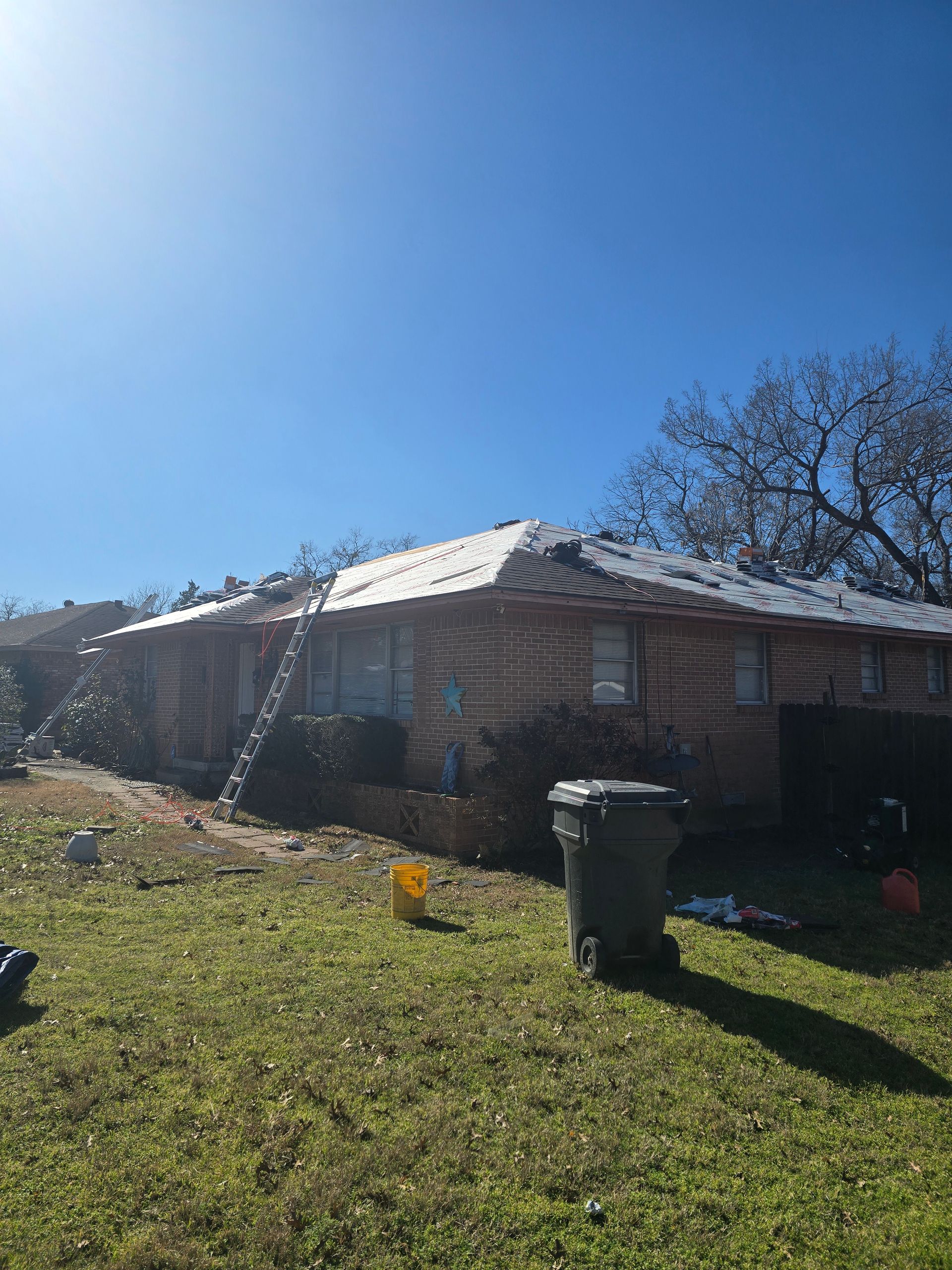 A brick building with a ladder leaning against it, roof partially covered in snow under a bright blue sky.