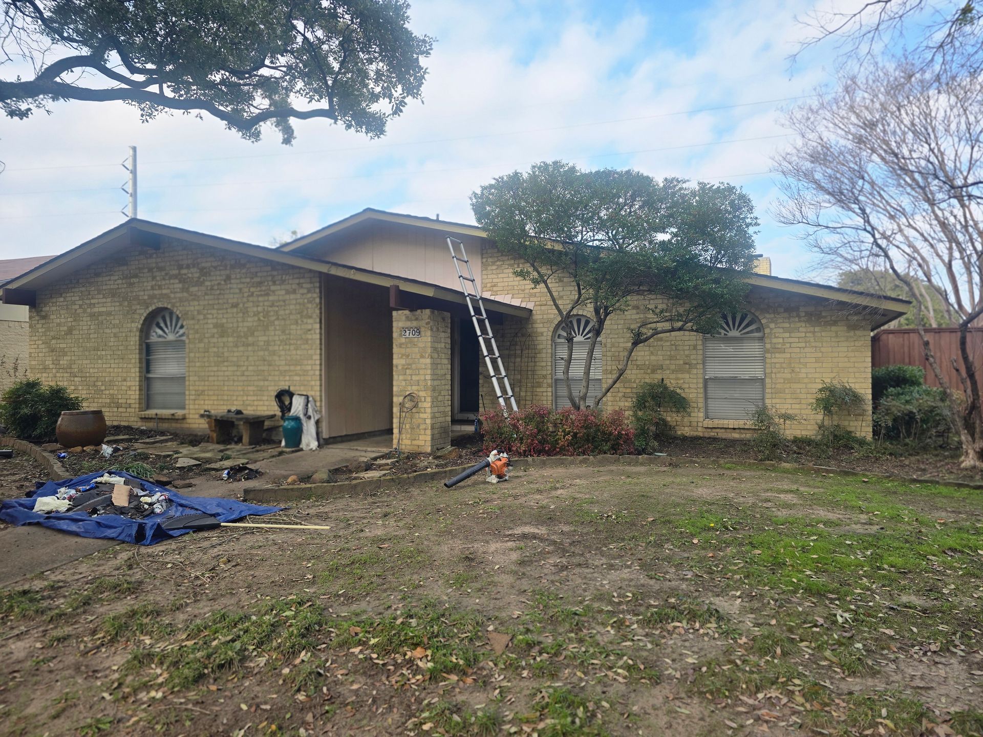 A one-story brick house with a ladder propped up near the entrance. Yard work in progress, tools scattered.