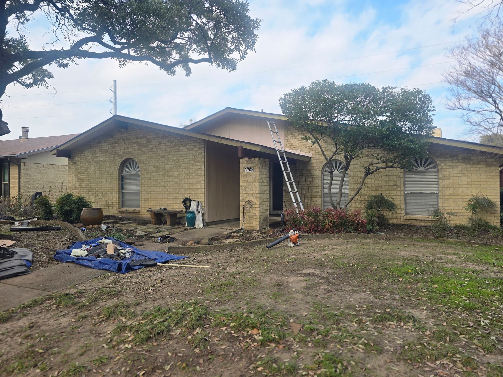 House exterior, yellow brick, with ladder leaning against the roof. Construction debris in the yard.