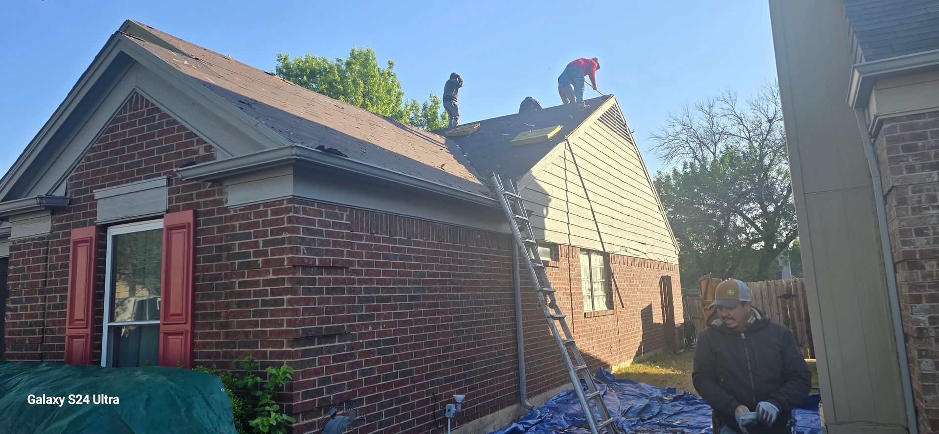Three workers on a brick house roof; one in red shirt, another at ladder.