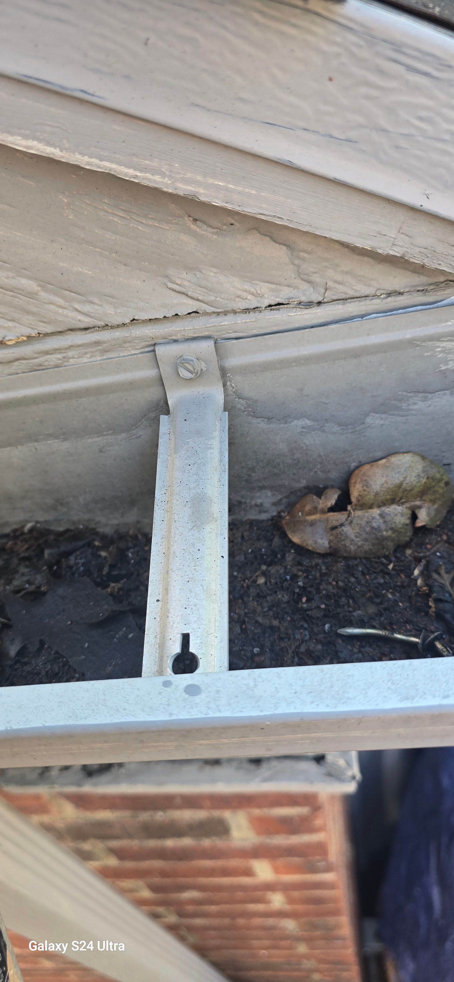 Gutter with debris, supported by metal bracket attached to a structure with brick exterior.