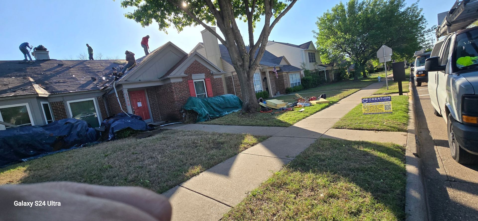 Workers on roofs of houses, grass and sidewalk in front, vans parked on street.