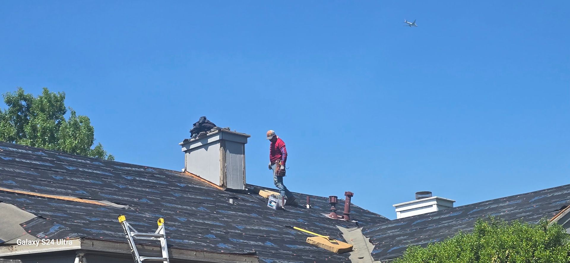 Two people working on a roof, blue sky, chimney, and a ladder are visible.