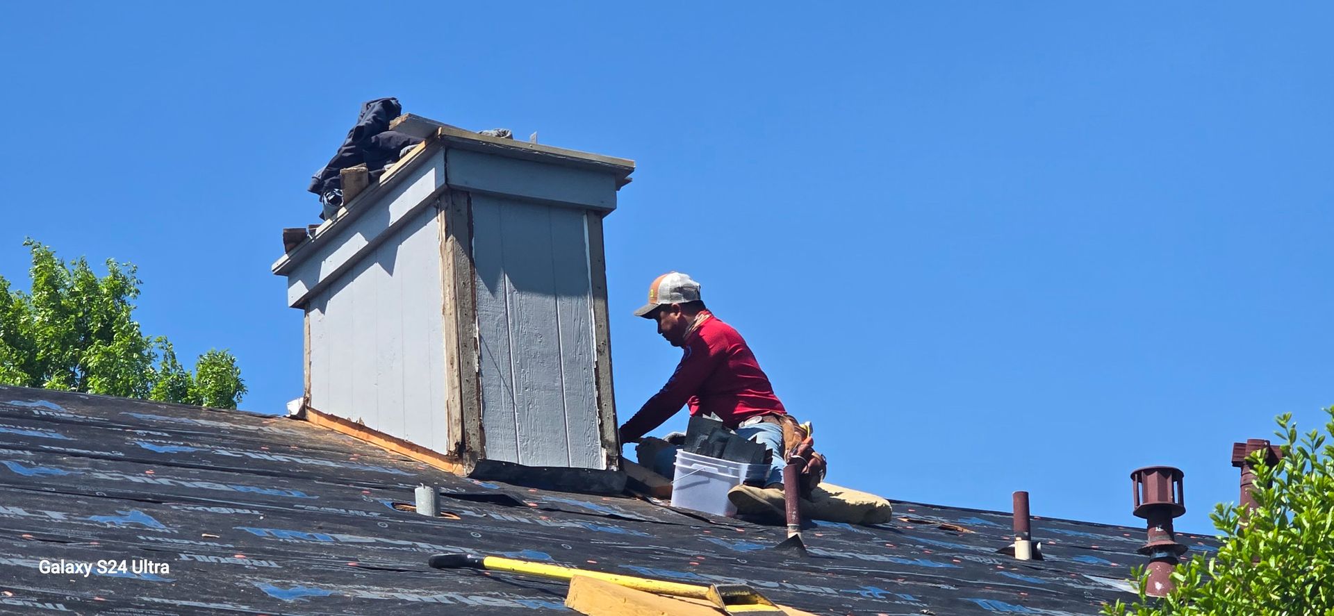Roofer repairing chimney on a black shingled roof under a blue sky.