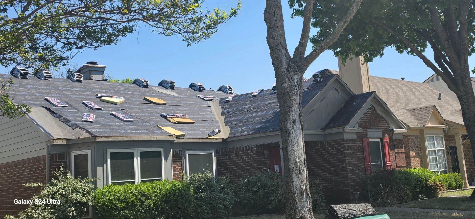 House with roof partially stripped of shingles, trees and blue sky visible.