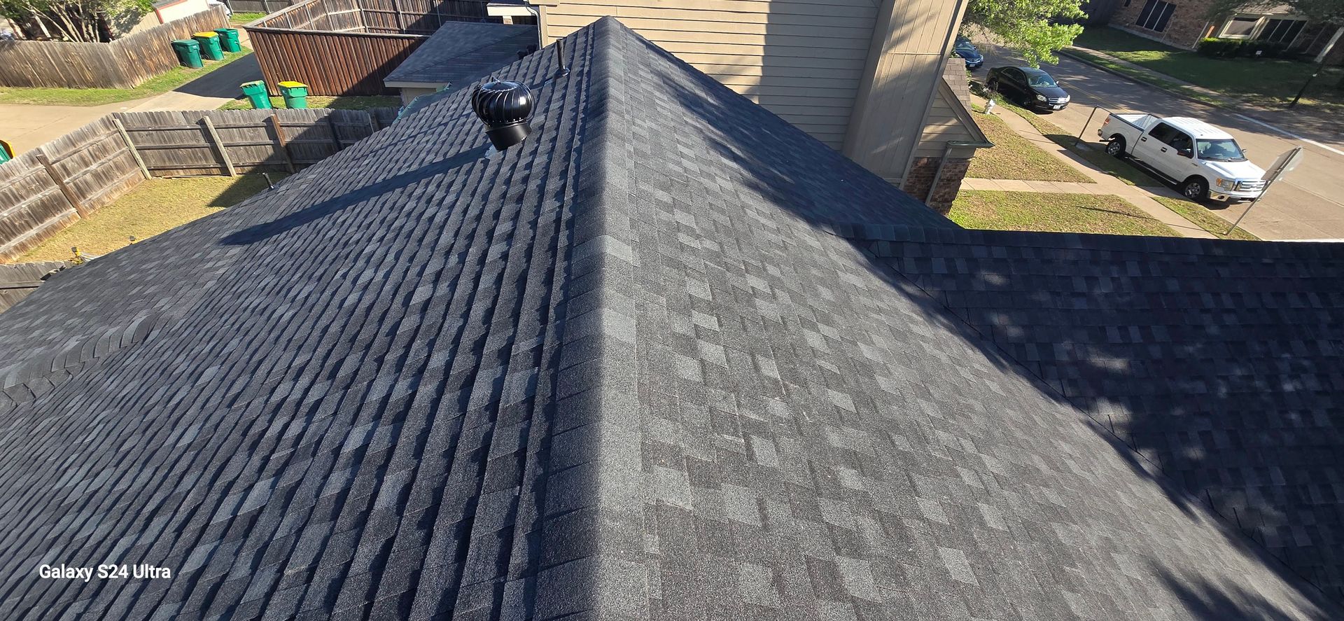 Overhead view of a dark roof with a person, grassy areas, a street with cars, and a fence.