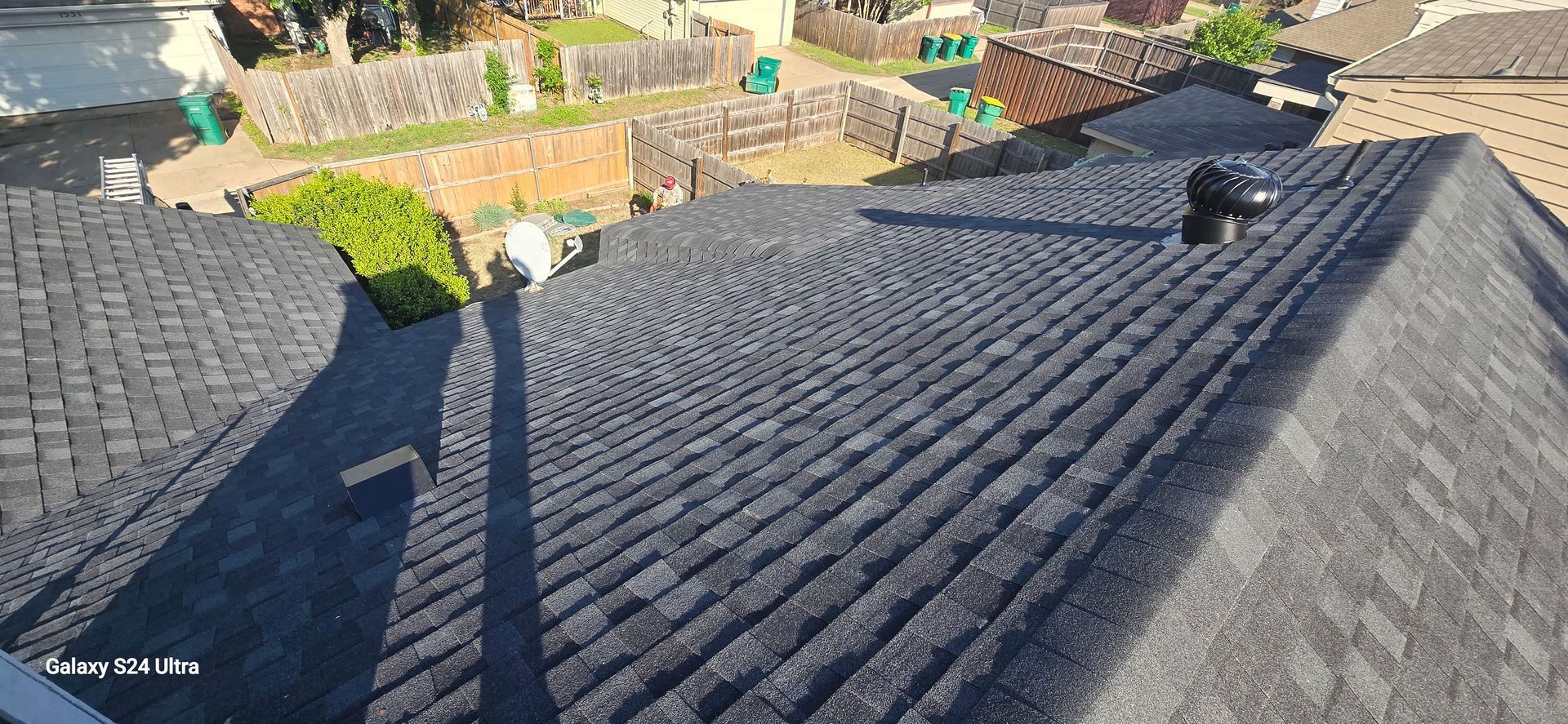 View of a dark shingled roof with a vent, surrounded by greenery and distant buildings.