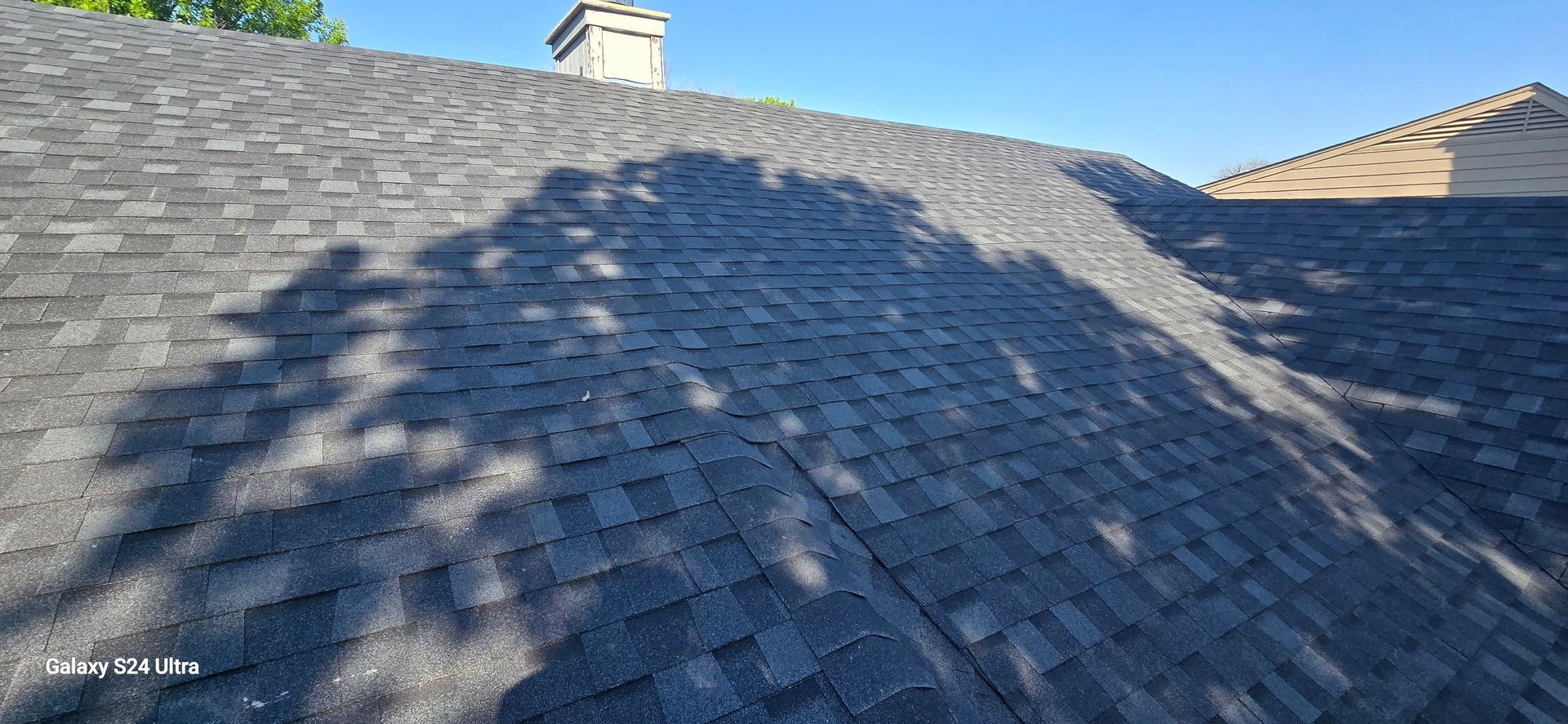 Dark gray asphalt shingle roof, with shadow of a tree and a chimney.