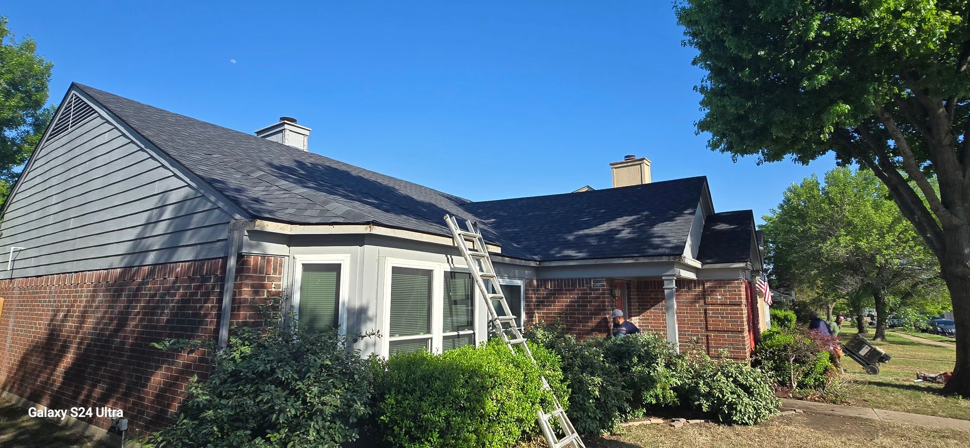 House with a partially replaced roof; brick exterior, ladder leaning against the side, clear blue sky.