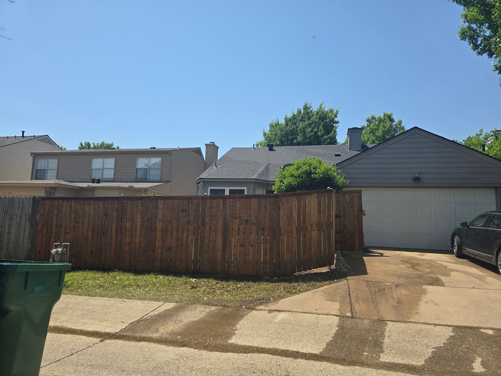Brown wooden fence surrounds a house with a gray garage door, a black car is parked nearby.