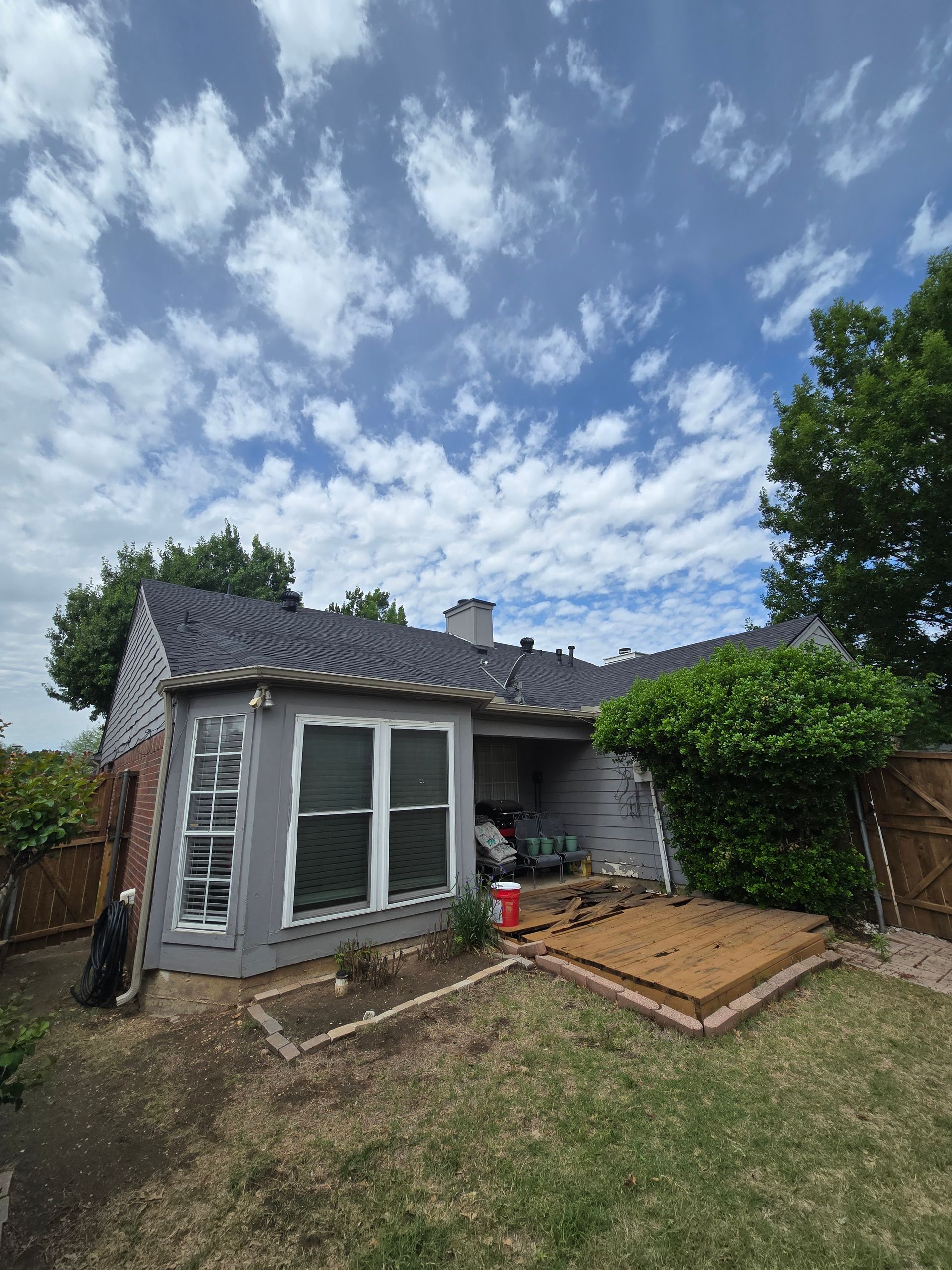 Gray house with a dark roof and a cloudy sky in a backyard. A small deck is in the yard.