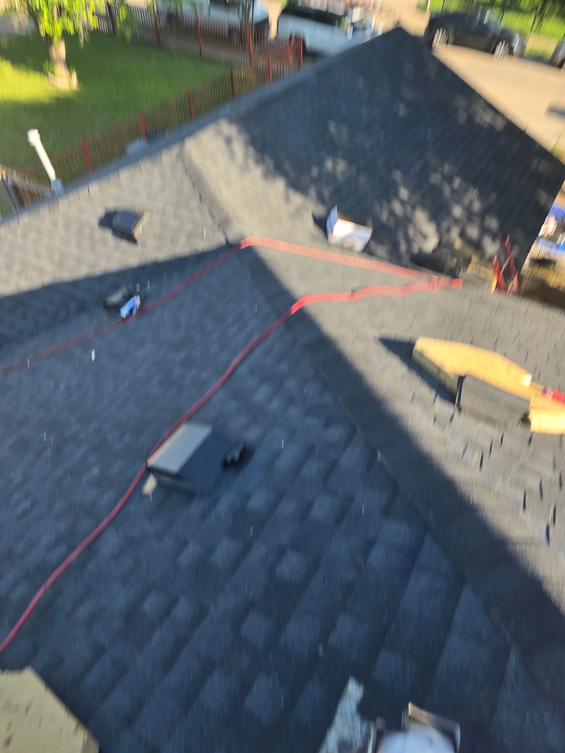 Rooftop with dark shingles; a red electrical cord lies across it, along with materials. Sunlight.