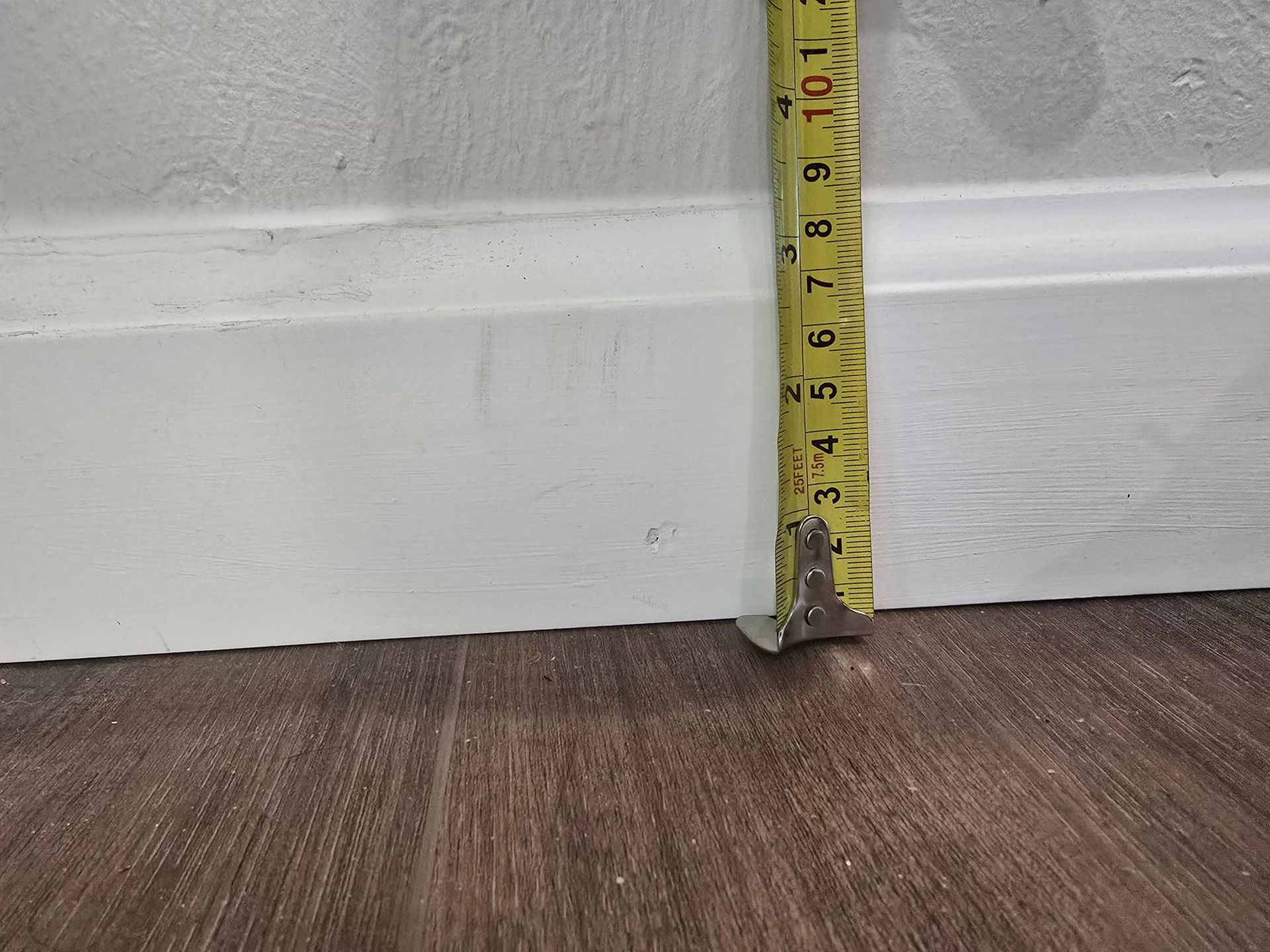 Yellow tape measure against white baseboard, wood floor. Measuring about 8 inches.