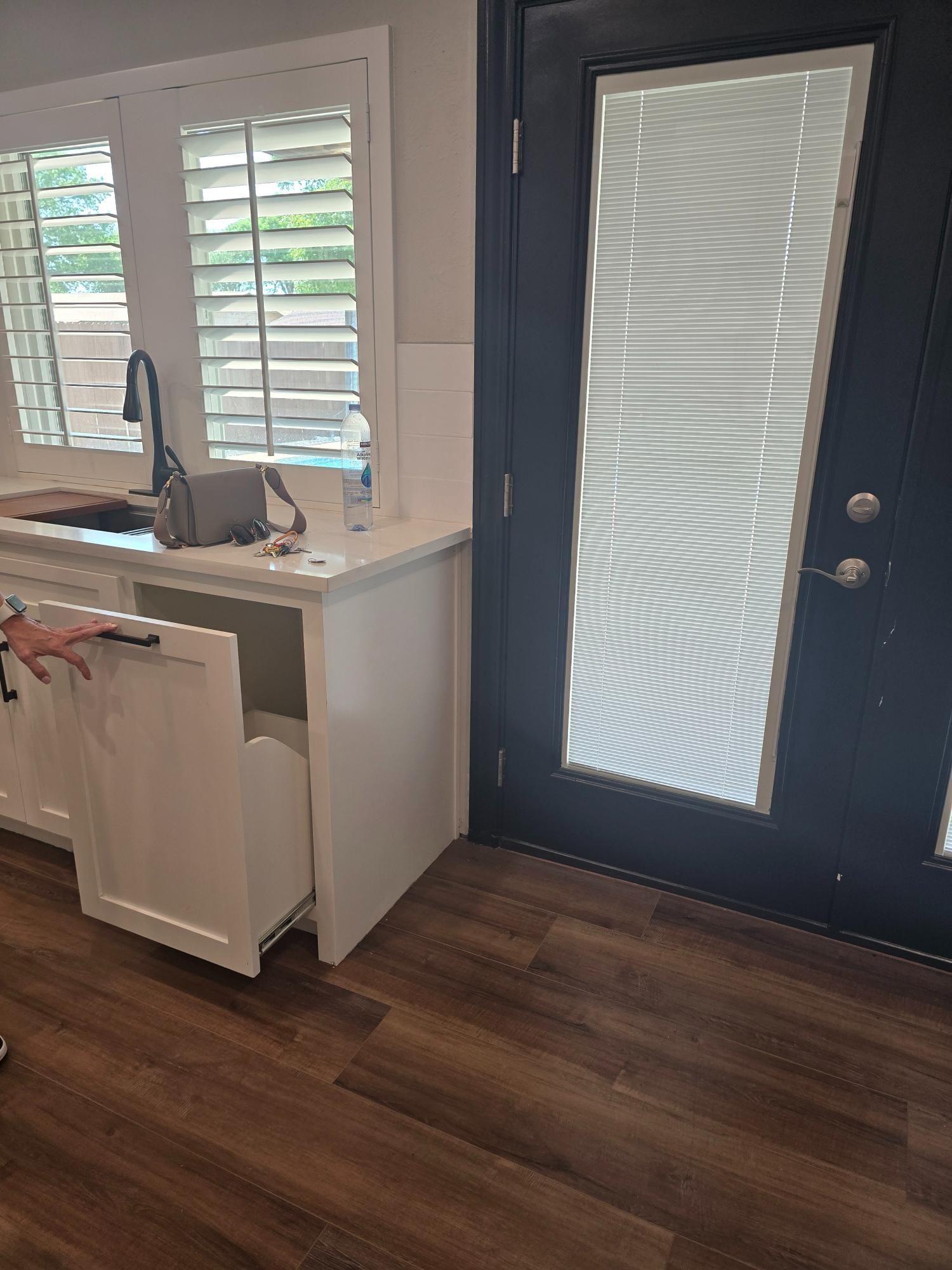 White kitchen cabinet with pull-out drawer open; black door with blinds next to it; dark wood floor.